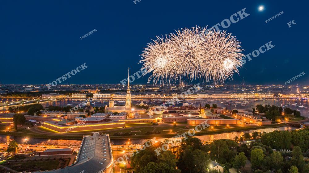 На фотографии изображён праздничный фейерверк над Петропавловской крепостью в Санкт-Петербурге. Ночной город освещён иллюминацией, подчеркивающей архитектуру крепости и окружающих мостов. На фоне видны река Нева и исторические здания города. Яркие вспышки фейерверка добавляют динамики этому впечатляющему пейзажу.